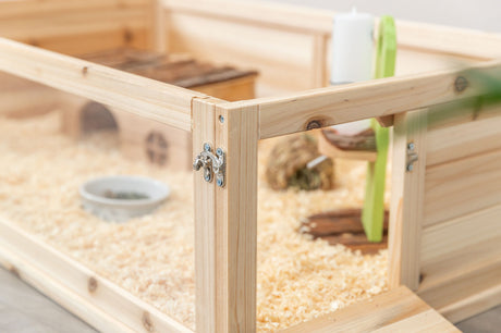 Holzgehege mit Transparentwand, innen Holzspäne, eine kleine Hütte, Futternapf und ein Spielzeug sichtbar.