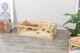 Holzterrarium auf Holzboden mit Einrichtungsgegenständen, enthält Einstreu, ein Röhrenhaus, Futternapf und Wasserflasche.