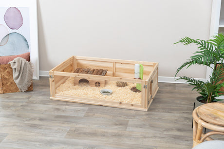 Holzterrarium auf Holzboden mit Einrichtungsgegenständen, enthält Einstreu, ein Röhrenhaus, Futternapf und Wasserflasche.