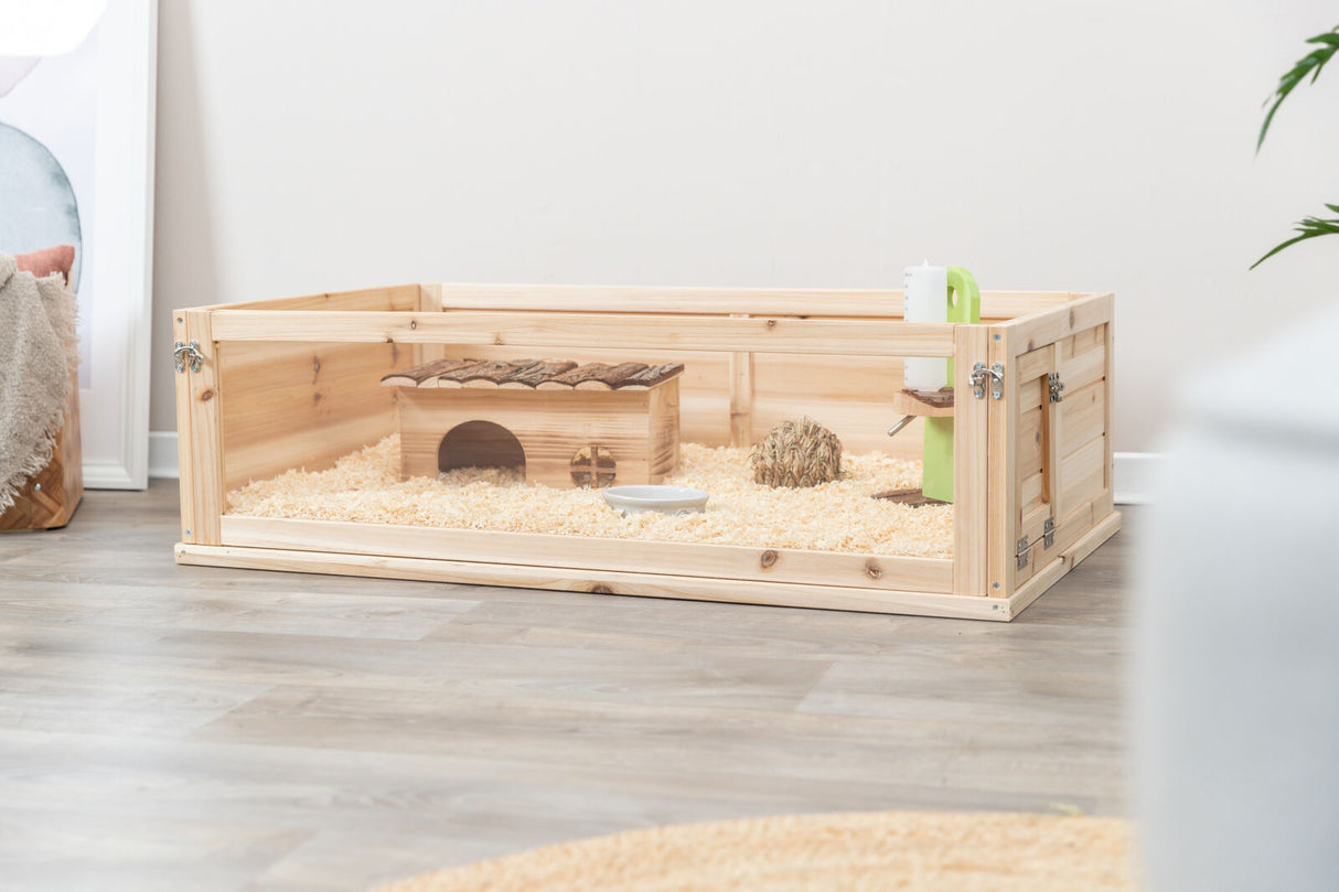 Holzgehege für Kleintiere mit Heu und Holzhäuschen auf Holzspänen, Trinkflasche an der Seite, Zimmerboden.