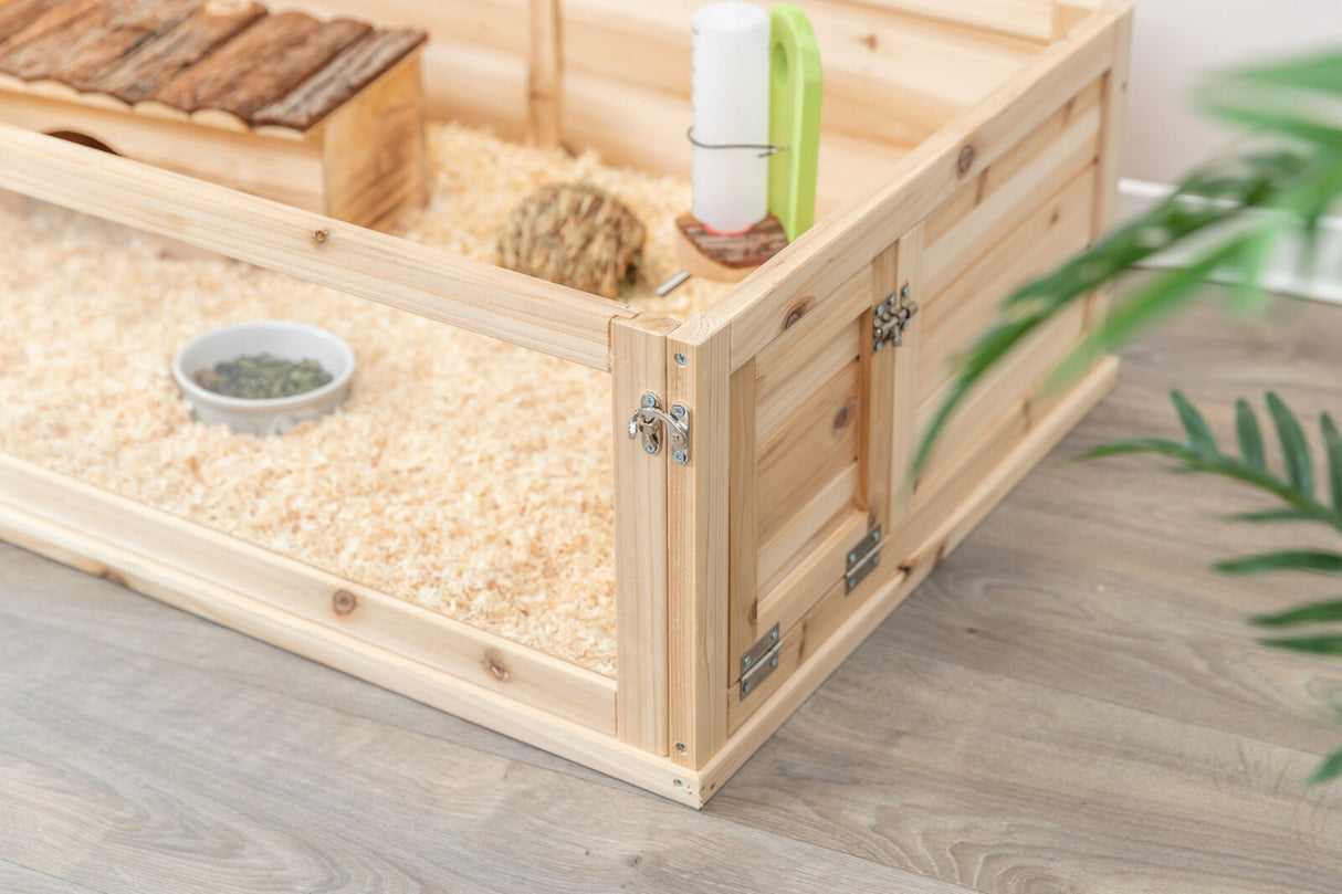 Holzgehege mit Spänen gefüllt, enthält eine kleine Hütte, einen Trinkbehälter, Heuballen und einen Napf.