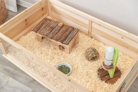 Holzgehege mit Einstreu, Holzhäuschen, Futter in einer kleinen Schale, Strohball und Wasserspender.