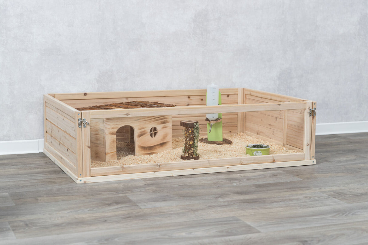 Holzgehege auf Holzfußboden mit Haus, Trinkflasche, Futtertrog und Späne als Einstreu.