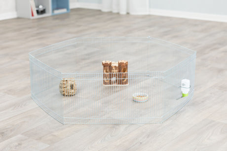 Metall-Laufstall auf Holzboden mit Futternapf, Wasserflasche, Holzspielzeug und knabberbarem Ball innen.