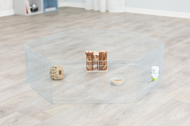 Metall-Laufstall auf Holzboden mit Futternapf, Wasserflasche, Holzspielzeug und knabberbarem Ball innen.