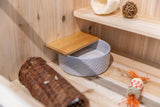 Graue Schale mit Sand, darüber ein Holzbrett, daneben ein geflochtenes Röhrchen und ein Spielzeug auf Holzregal.