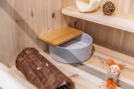 Graue Schale mit Sand, darüber ein Holzbrett, daneben ein geflochtenes Röhrchen und ein Spielzeug auf Holzregal.