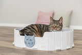Katze liegt in einem runden, hellen Bett mit Katzenmotiv im Vordergrund.