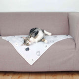 Katze liegt auf einer weißen Decke mit Katzenmotiven auf einem grauen Sofa, der Holzboden ist sichtbar.