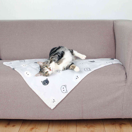 Katze liegt auf einer weißen Decke mit Katzenmotiven auf einem grauen Sofa, der Holzboden ist sichtbar.