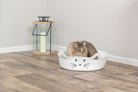 Eine Katze liegt in einem weißen Korb mit Katzengesicht auf einem Holzboden, neben einer Laterne.