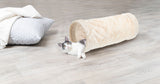 Weiß-graue Katze liegt in beige-rohrförmigem Kissen auf hellem Holzboden, neben einem weißen Kissen.