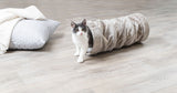 Eine schwarz-weiße Katze schaut aus einer flauschigen, grauen Röhre auf einem Holzfußboden, daneben ein graues Kissen.