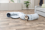 Eine Katze sitzt in einem flauschigen, grauen Tunnel auf einem Holzfußboden im Wohnzimmer.