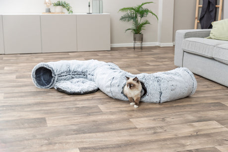 Eine Katze sitzt in einem flauschigen, grauen Tunnel auf einem Holzfußboden im Wohnzimmer.