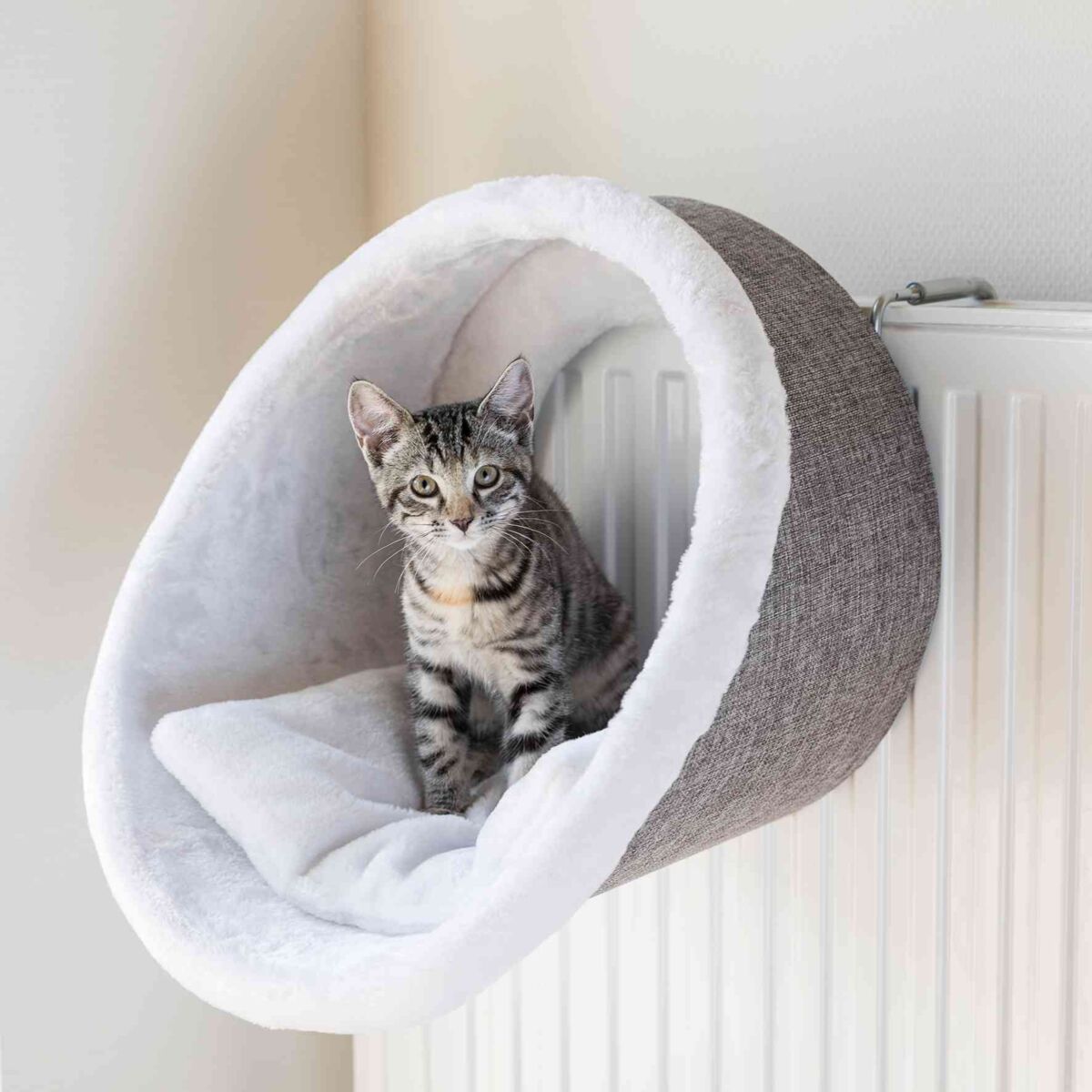 Eine graue Katze sitzt in einer hellgrauen, an der Heizung hängenden Höhle mit weißer Innenpolsterung.