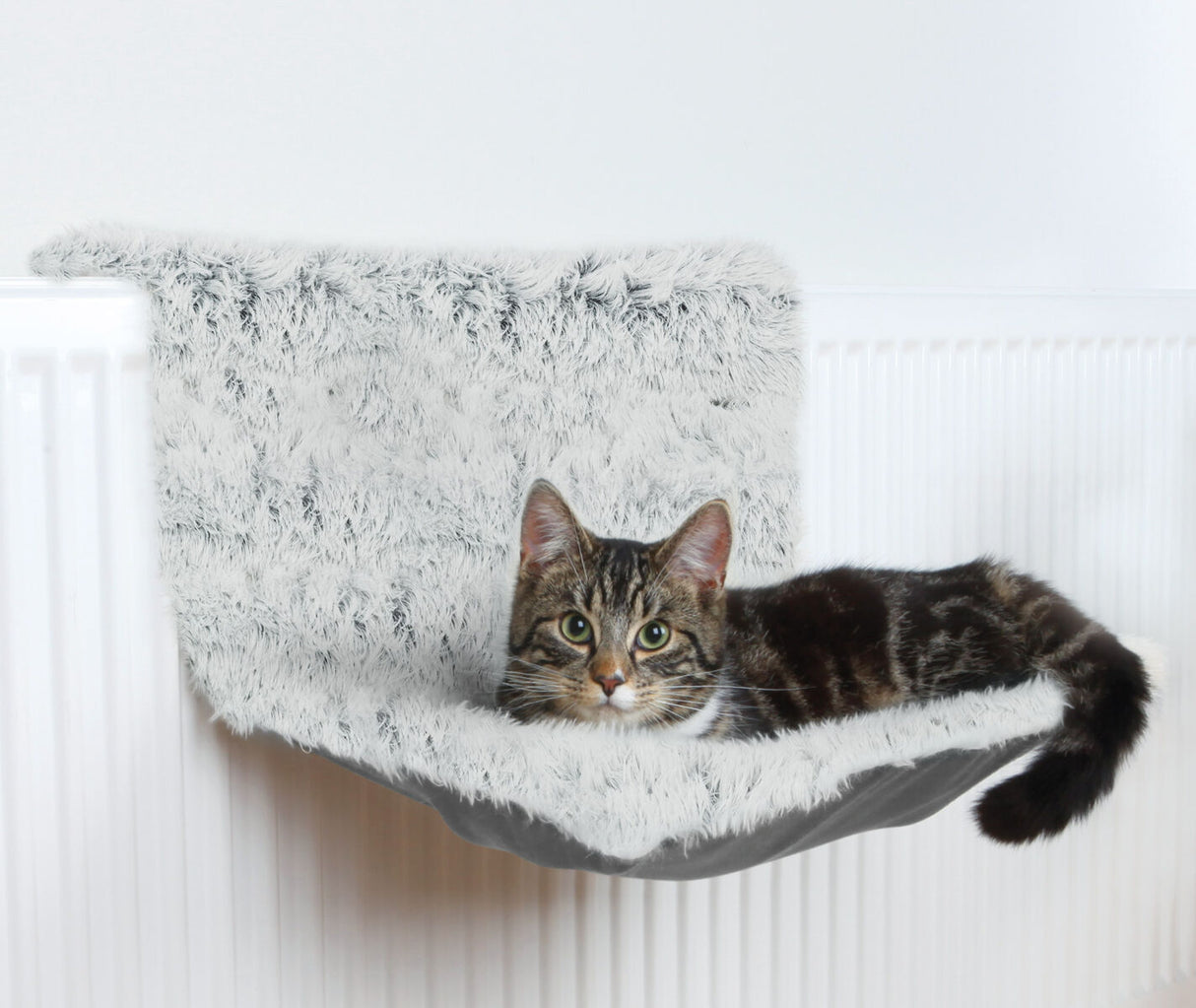 Eine getigerte Katze liegt auf einer grauen, pelzigen Hängematte, die an einem weißen Heizkörper angebracht ist.