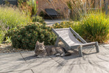 Eine getigerte Katze liegt neben einem kleinen Liegestuhl auf einem gepflasterten Gartenweg.