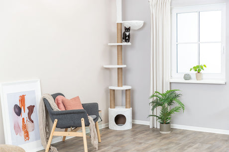 Wohnzimmer mit modernem Kratzbaum, Katze auf mittlerer Plattform, Sessel und Pflanzen, großes Fenster rechts.