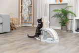 Eine schwarze Katze mit weißen Pfoten spielt an einem Kratzbaum in einem Wohnzimmer mit Holzfußboden.