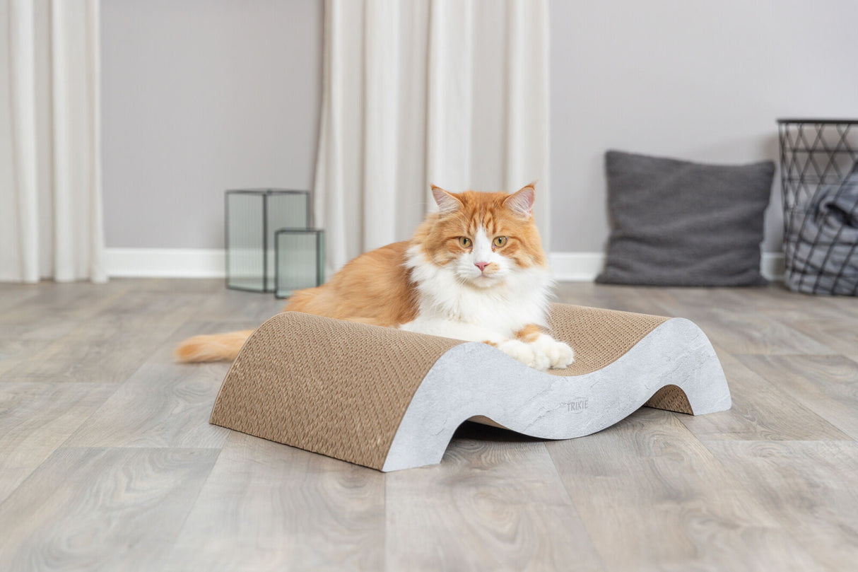 Eine getigerte Katze ruht auf einem wellenförmigen Kratzbrett aus Pappe auf einem Holzfußboden in einem hell eingerichteten Raum.