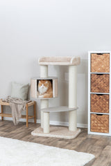 Katze sitzt in einer beigen Kratzbaumhöhle neben einem Holzhocker und einem Regal mit Körben.