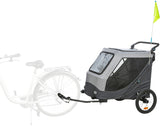 Grauer Fahrradanhänger mit zwei Rädern und gelber Fahne, an einem Fahrrad befestigt.
