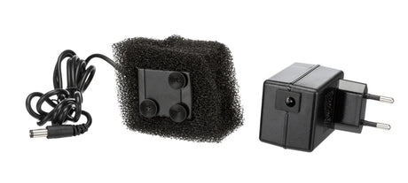 Schwarzes Netzteil mit europäischem Stecker und Kabel neben rechteckigem Schaumstoffstück mit Saugnäpfen.