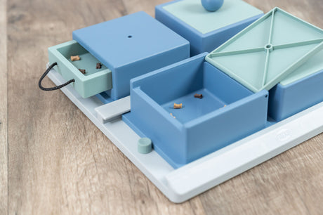 Ein blau-grauer Intelligenzspielzeug mit mehreren Fächern und Schubladen, teilweise offen, auf einer Holzoberfläche.