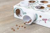Papierbox mit mehreren Öffnungen und Röhren, aus denen braune Stückchen auf einen Holzboden fallen.