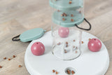 Rundes weißes Spielzeug mit rosa Kugeln und braunen Leckerlis auf einem Holzboden, zylinderförmige, transparente Säule in der Mitte.
