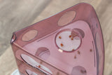 Transparenter, rosa Behälter mit runden Öffnungen, innen liegen kleine braune Stücke auf einer weißen Fläche.