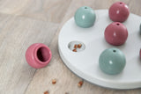 Rundes Spielbrett mit rosa und blauen Kugeln auf Holzboden, sichtbare Leckerli in einer Öffnung.