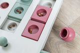 Spielbrett in Blau, Rosa und Weiß mit runden Vertiefungen und Leckerli, auf Holzfußboden liegend, rosa Hütchen daneben.