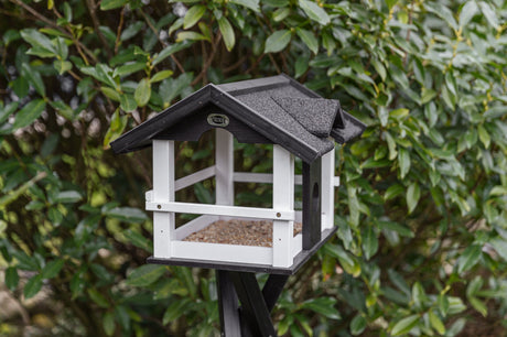 Vogelhaus auf einem Ständer in einem Garten, mit schwarzem Dach und weißen Wänden, umgeben von grünen Blättern.
