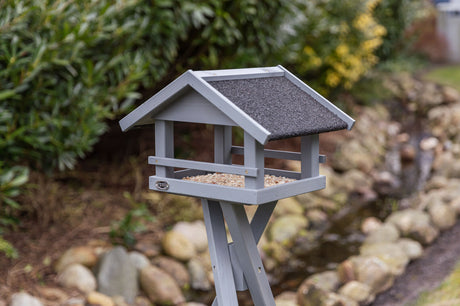 Graues hölzernes Vogelhaus mit schwarzem Dach auf einem Ständer, im Garten positioniert, mit Hintergrund aus Büschen und Steinen.