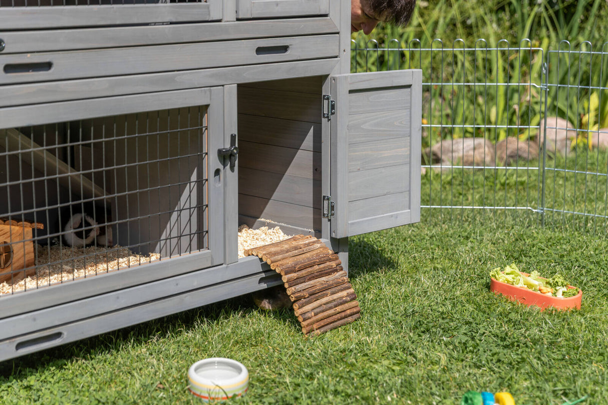 Graues Kleintiergehege mit offener Tür steht auf Gras, neben einem Zaun, Futternapf und Spielzeug.