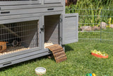 Graues Kleintiergehege mit offener Tür steht auf Gras, neben einem Zaun, Futternapf und Spielzeug.
