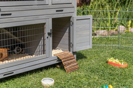 Graues Kleintiergehege mit offener Tür steht auf Gras, neben einem Zaun, Futternapf und Spielzeug.