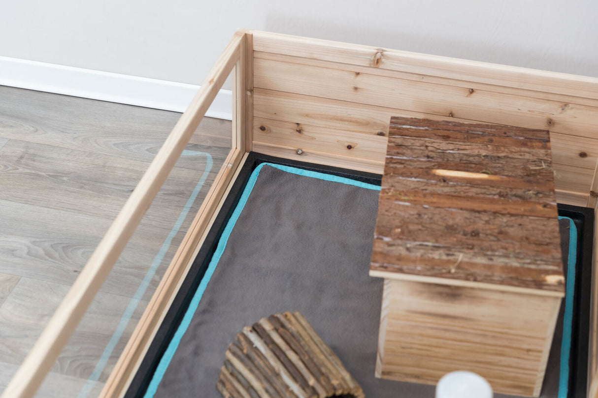 Holzkäfig mit grauer Matte und hölzernem Haus auf Laminatboden, Seitenwände aus Glas.