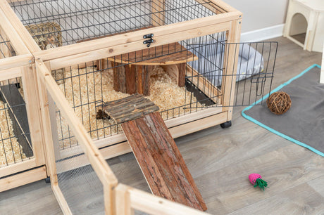 Holzkäfig mit offenem Gittertürchen, Holzrampe und Späne auf dem Boden, Spielzeug auf heller Holzfläche.