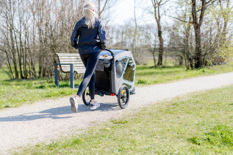 Eine Person in Sportkleidung joggt mit einem Fahrradanhänger auf einem Kiesweg in einem Park.