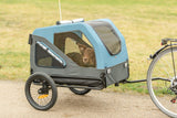 Hund sitzt in blauem Fahrradanhänger neben angekoppeltem Fahrrad auf einem Weg.