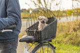 Kleiner Hund sitzt in einem schwarzen Fahrradkorb mit Gitter auf einem Fahrrad im Freien.