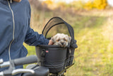 Kleiner Hund sitzt in einer Fahrradbox mit Abdeckung, gehalten von Person in blauer Jacke.