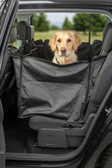 Ein Hund sitzt in einem schwarzen Kofferraumschutz im Auto, umgeben von grüner Landschaft.
