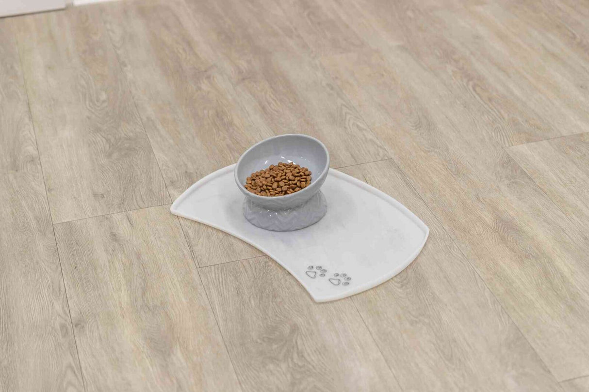 Graue Futterschale mit Trockenfutter auf weißer Matte mit Pfotensymbolen auf hellem Holzboden.