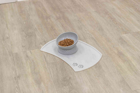 Graue Futterschale mit Trockenfutter auf weißer Matte mit Pfotensymbolen auf hellem Holzboden.