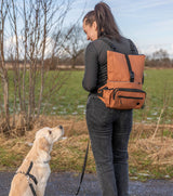 Frau mit braunem Rucksack und schwarzem Shirt steht auf dem Pfad neben einem sitzenden Hund, der sie anschaut.