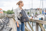 Eine Person steht auf einem Holzsteg neben einem Hafengelände mit Segelbooten, einen schwarzen Rucksack tragend, aus dem ein kleiner Hund schaut.